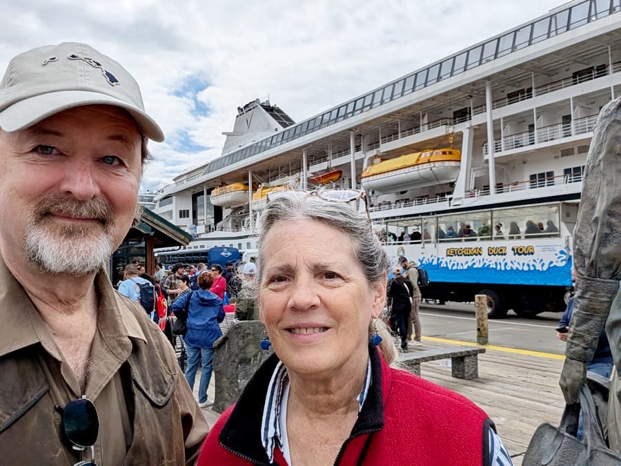 Randy and Kit smile for a selfie at a busy dock with a large cruise ship and a sightseeing bus in the background. Other people are gathered around, and the sky is partly cloudy.