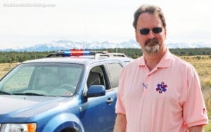 A man in a pink polo shirt with an EMS logo stands in front of a blue emergency vehicle with flashing lights against a backdrop of snow-capped mountains.
