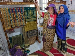 Two women dressed in Indonesian woven textiles smile in a fabric shop with colorful cloths and a Dekranasda Anambas sign overhead.