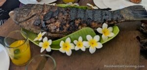 A whole grilled fish on a large leaf, topped with yellow and white plumeria flowers, drinks and plates on a wooden table.