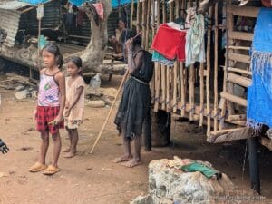 Two young girls and an elderly woman stand by a rustic house with laundry hanging; another woman sits inside. Dirt, stones, and makeshift structures outside.