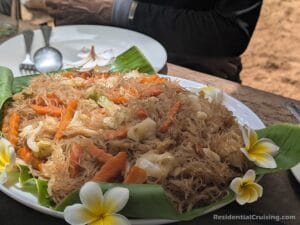 Stir-fried glass noodles with veggies and frangipani flowers on banana leaves; person in black behind, utensils on the table.