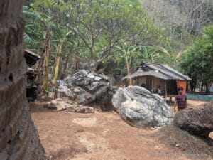 A rural scene with large rocks, a thatched hut, and lush tropical plants. A person in stripes walks near the hut amid greenery.