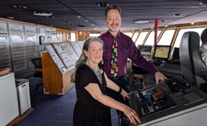 Kit wearing a black dress standing at a console of the very spacious ship's bridge. There is equipment on the back wall, screens showing maps and information, and the duty officer sitting in the captain's chair. Randy is next to Kit, smiling, wearing a purple shirt and a colorful tie of "Looney Tunes" cartoon characters.