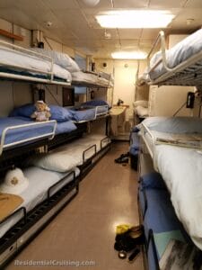 Narrow ship crew cabin with stacked bunk beds on both sides, blue bedding, personal items like shoes and a teddy bear, and a white sailor hat. A tight corridor runs down the middle.