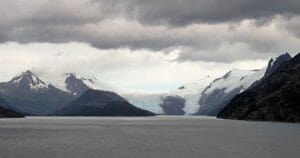 A wide, calm sea stretches toward snow-covered mountains under a cloudy sky, with a large glacier visible between the peaks in the distance.