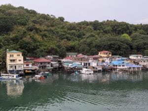 Bright stilt houses line a calm waterfront with docked boats; green forested hills rise behind the village under a gray sky.