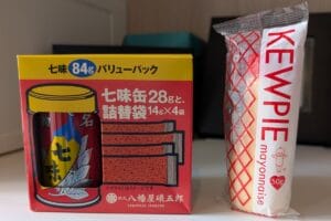 A box of Japanese shichimi spice mix with a red and yellow design sits next to a small tube of Kewpie mayonnaise with a diamond-patterned wrapper. Both are on a light-colored surface.