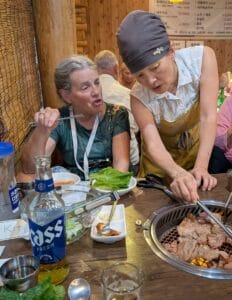 A woman in an apron is grilling meat at a restaurant table while Kit speaks. Plates of food, a bottle of beer, and vegetables are on the table. Other diners are in the background.