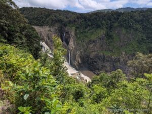 A tall, narrow waterfall cascades down a rocky cliff surrounded by dense green forest and shrubbery, with mountains and a partly cloudy sky in the background.