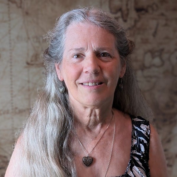 An older woman with long gray hair, patterned sleeveless top, heart earrings, and pendant necklace smiles before a sepia map backdrop.