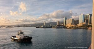 From aboard ship with clear flat water showing, we see the "Aloha Tower" ashore in Honolulu, along with buildings and shoreline. A tugboat near the ship is ready to jump to action as needed to nudge the ship in the right direction. It is near sunset, with a warm light, and puffy clouds partially obscuring a blue sky.