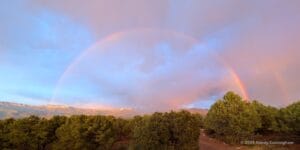 A beautiful blue Colorado sky obscured by scattered clouds. It is raining in the distance, and we can see the full arches of a double rainbow from edge to edge.