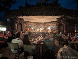 It's evening, and a crowd sitting at tables are looking up at a covered outdoor stage where dancers are twirling lit torches while wearing bright island costumes.