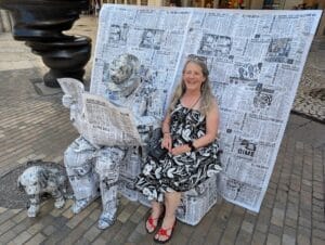 Kit in a black and white dress sits laughing on a bench next to a street performer and dog statue covered in newspaper print, with a matching newspaper-print backdrop behind them.
