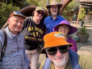 Five smiling adults wearing colorful hats and sunglasses pose together outdoors on a sunny day, with greenery and part of a large sculpture in the background.