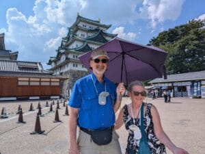 Randy and Kit smile under a purple umbrella in front of Nagoya Castle in Japan. Both wear sunglasses and battery-powered fans around their necks. The sunny day casts clear shadows, and other visitors are visible in the background.
