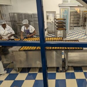 Looking through a window into a kitchen area where two workers are at a steel table holding around eight trays, each filled with tiny custard-filled pastries coming out of the oven. There are multiple cooling racks in the background, one has several more full trays.