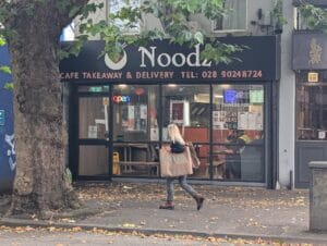 A glass window storefront with a sign above reading, "Noodz — Cafe Takeaway & Delivery". Tables are in view, with people sitting there, though mostly people just pick up things to bring home. Super good, and reasonable prices, especially for Belfast.