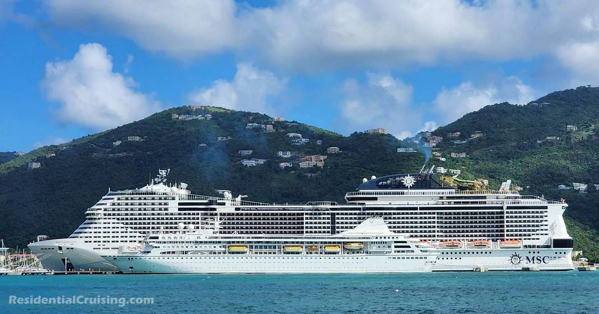Two white cruise ships: one is the gigantic MSC Virtuoso, looming over the tiny Villa Vie Odyssey. They are docked side by side in a turquoise harbor, with green, hilly islands and scattered white houses in the background under a partly cloudy sky.