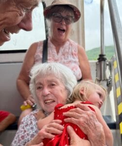 A joyful elderly woman hugs a young girl in a red dress. Behind them, two smiling adults watch, one wearing glasses and a sunhat. The scene appears to be on a bus or tram with outdoor light in the background.