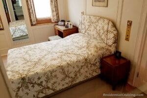 Aboard ship, a neatly made bed with a floral-patterned bedspread and matching padded headboard sits between a wooden nightstand and a small desk with a stool in a cozy, well-lit bedroom with cream-colored walls.