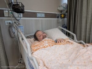 A man with a beard lies in a hospital bed, covered by a patterned blanket. He smiles slightly, surrounded by medical equipment in a hospital room marked “RR 10.”.