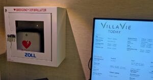 A box labeled EMERGENCY DEFIBRILLATOR is mounted on a wall next to a digital sign displaying services and hours for VILLA VIE TODAY.