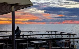 A young woman stands on the deck overlooking a calm sea at sunset, with vibrant orange, pink, and purple clouds filling the sky. Rooftop and railings frame the scenic ocean view.