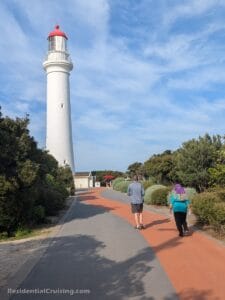 Two people walk along a curved path near a tall white lighthouse surrounded by greenery on a sunny day. The sky is blue with scattered clouds.