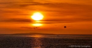 Sunset over the ocean with an orange sky, a distant island, and a parasailor above the water. Gentle waves are visible, and the scene is calm and serene.