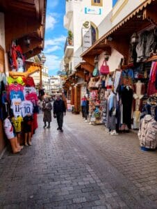 A market atmosphere on a narrow, pedestrian, brick street. Colorful T-shirts dominate the left and clothing and bags on the right, while a local couple are strolling toward the camera.