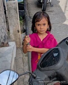 A young girl in a bright pink top stands on a street, holding a metal pole and looking at the camera. A motorcycle is parked in the foreground.