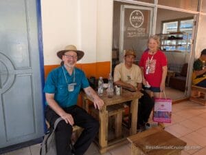 Three people smile at a small wooden table by an orange and white wall; two seated, one standing with a pink shopping bag.