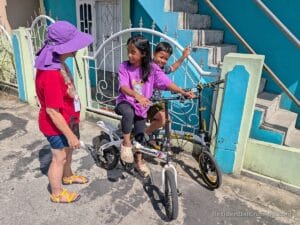 A woman in a purple hat and red shirt stands beside two smiling kids on bikes, with a blue building and stairs in the background.