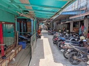A narrow alleyway with motorcycles parked on both sides, teal awnings, weathered buildings, open doors, and clothes hanging.