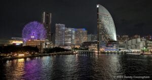 Night view of the Yokohama city skyline with illuminated buildings, a large ferris wheel glowing purple, and their reflections shimmering on the water in the foreground.