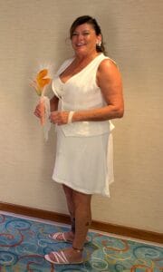 A smiling woman stands indoors wearing a sleeveless white dress and matching vest, holding a bouquet with orange flowers. She has earrings, a bracelet, sandals, and a tattoo on her left leg. The carpet is patterned and colorful.