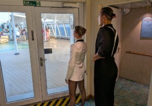 Two performers in formal dance attire stand inside by a glass door, looking out at a deck where people are gathered. The exit sign is above the door, and the indoor carpet contrasts with the outdoor deck.