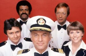 Five people in formal white maritime uniforms pose in front of a bright red background. The man in the center wears a captains hat, while the others stand around him, smiling and looking at the camera.