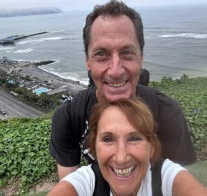 A smiling man and woman take a selfie on a grassy hill overlooking the ocean, coastal road, beach, and distant pier on a hazy day.