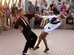 Two dancers in vintage-style outfits energetically perform on a deck as a large, seated audience watches and applauds. The man wears black with suspenders, and the woman wears a white suit with black accents.