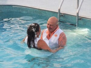 A man and woman, dressed in formal clothes, stand close together in a swimming pool, looking at each other and smiling. The woman has flowers in her hair, and the man is wearing an orange shirt with a white vest.